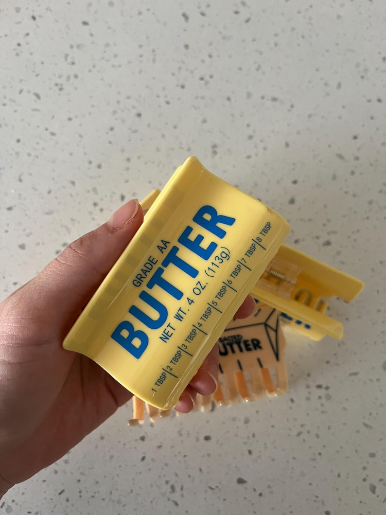 May include: A yellow plastic hair claw clip designed to resemble a stick of butter. The clip features blue text that reads GRADE AA BUTTER, NET WT. 4 OZ. (113g), and includes measurement markings from 1 TBSP to 8 TBSP along the side of the clip.