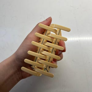 May include: A pale yellow hair claw clip featuring a series of parallel teeth on both sides. The surface of the clip is decorated with thin, black geometric line patterns that form a repeating angular design across the light-colored plastic body.
