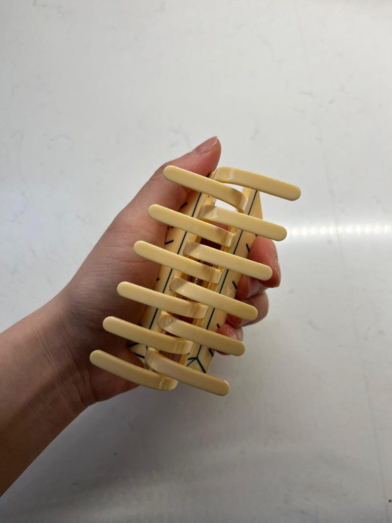May include: A pale yellow hair claw clip featuring a series of parallel teeth on both sides. The surface of the clip is decorated with thin, black geometric line patterns that form a repeating angular design across the light-colored plastic body.