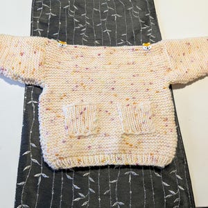 May include: A hand-knitted, speckled sweater in cream, yellow, and purple. The sweater has two front pockets and small animal-shaped buttons on the shoulders. The garment is displayed on a dark grey patterned surface.