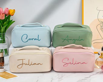 May include: Four personalized makeup bags in different colors. The bags are light blue, green, beige, and pink. Each bag has a gold zipper and a gold handle. The bags are embroidered with the names "Coral", "Angela", "Julian", and "Selina".