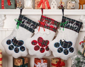 May include: Three white Christmas stockings shaped like a dog's paw, each with a plaid cuff and the names "Charlie", "Malvina", and "Bailey". The paw pads are embroidered with plaid fabric. Green and red ribbons add festive accents.