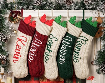 May include: Five cable-knit Christmas stockings with names embroidered in white. The stockings are cream, red, and green, with reindeer antlers at the top. Festive holiday decor.