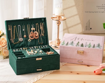 Puede incluir: Una caja de joyería verde oscuro abierta, con varios collares, aretes y anillos. Una caja de joyería rosa cerrada con el nombre "Athena" y un diseño de bosque está al lado. Un candelabro dorado está en el fondo.