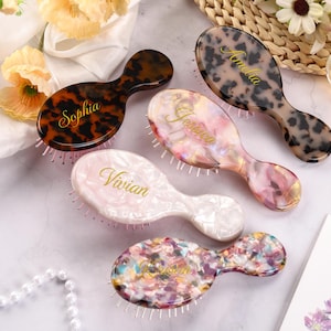May include: Five personalized hairbrushes in various colors and patterns, including tortoiseshell, pink, and multicolored designs. Each brush features a different name in gold script: Sophia, Amelia, Jessica, Vivian, and another name. The brushes are arranged on a light-colored surface.