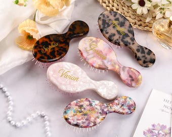 May include: Five personalized hairbrushes in various colors and patterns, including tortoiseshell, pink, and multicolored designs. Each brush features a different name in gold script: Sophia, Amelia, Jessica, Vivian, and another name. The brushes are arranged on a light-colored surface.