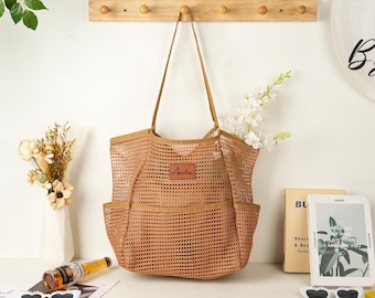 May include: A tan mesh tote bag with two front pockets and long handles. A small brown leather patch with the name "Amelia" is sewn on the front. The bag is displayed with flowers, books, and a bottle.