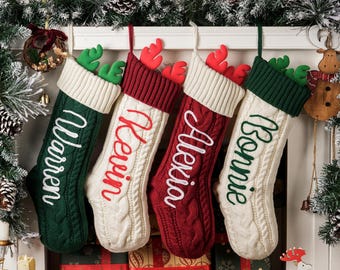 May include: Four cable-knit Christmas stockings in green, red, and cream, each with a name embroidered in a contrasting color. The stockings are decorated with antler accents and hang from a white mantelpiece.
