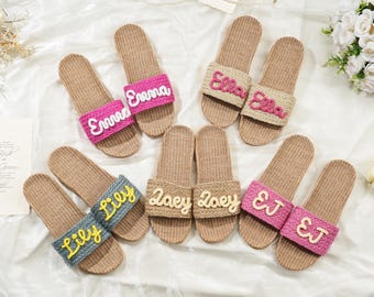 May include: Five pairs of slip-on sandals with woven tan jute footbeds and textured straps. The straps feature embroidered names including Emma, Ella, Lily, Zoey, and EJ in various colors like pink, yellow, and white, arranged on a white fabric.