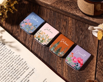 May include: Four floral-themed magnetic bookmarks with names: Sophia, Julian, Salome, and Carroll. Each bookmark features a different color and floral design, including pink, orange, and blue. The bookmarks are arranged on a dark wooden surface.