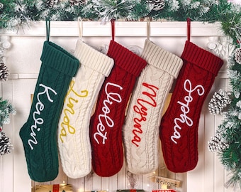 May include: Five personalized Christmas stockings in green, cream, and red, each with a different name embroidered in a contrasting color. The stockings are hanging against a white background, decorated with pine branches and pine cones.