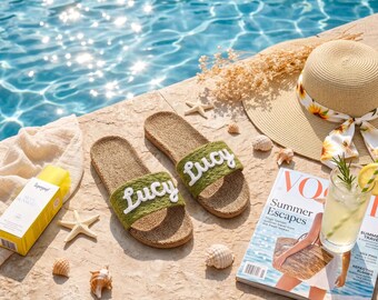 May include: A pair of olive green slides with the name Lucy embroidered in white script on the strap. The slides feature a textured tan footbed and are displayed alongside a straw hat with a sunflower ribbon, a magazine, and sunscreen.