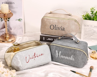 May include: Three glittery cosmetic bags in beige, silver, and gray, each with a handle and a zipper. The bags are personalized with the names "Olivia," "Victoria," and "Hannah" in elegant script. The bags are open, revealing the interior.