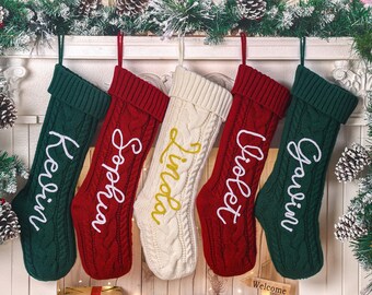 May include: Five cable-knit Christmas stockings in green, red, and cream, each with a name embroidered in a contrasting color. The stockings are hanging from a white mantelpiece decorated with pine boughs and pinecones.