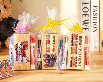 May include: Three gift sets for teachers, each wrapped in clear cellophane and tied with a ribbon. The sets include wooden blocks with teacher-themed graphics and text, along with dry-erase markers. One block reads "Mrs. Elly."