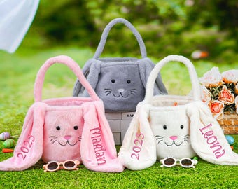 May include: Three plush bunny-shaped Easter baskets with personalized names. The baskets are pink, white, and gray. The pink basket has the name "Makayla" embroidered on it, the white basket has the name "Leona" embroidered on it, and the gray basket has the year "2023" embroidered on it.