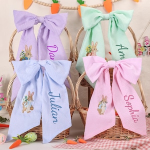 Puede incluir: Cuatro lazos decorativos de Pascua en cestas. Cada lazo es de un color pastel diferente: lila, verde menta, azul claro y rosa. Cada lazo tiene un nombre bordado y una imagen de conejito. Los lazos son a rayas.