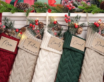 May include: Five cable-knit Christmas stockings in red, tan, cream, green, and tan, each with a personalized leather tag. The tags are inscribed with names like "Mom" and "Dad". The stockings are decorated with pine boughs, berries, and pinecones.
