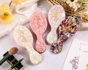 May include: Four oval-shaped hairbrushes in various colors: white, pink, and a multicolored design. Each brush has a handle and a name in gold script: Kristin, Amelia, Sophia, and a multicolored brush. Makeup brushes are in the foreground.