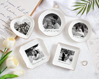 May include: A collection of white ceramic trinket dishes in various shapes, each featuring a black and white photograph and gold trim. The dishes include a heart, round, and square shapes, with handwritten text.