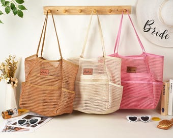 May include: Three mesh tote bags in tan, cream, and pink, each with a front pocket and a leatherette name tag. The bags have long handles and are suitable for beach or shopping.