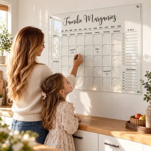 Könnte beinhalten: Ein wandmontierter Monatskalender aus klarem Acryl mit der Aufschrift Familie Marganiez. Das Raster enthält Bereiche für Notizen, To-Do-Listen und Menü-Ideen, wobei die Wochentage und Monate auf Deutsch beschriftet sind.