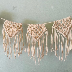 May include: Cream macrame banner featuring triangular shapes with detailed knotting and long fringe. The banner is suspended against a light blue wall, creating a bohemian aesthetic. The banner is suitable for home decor.