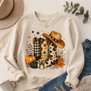 May include: A beige crewneck sweatshirt featuring a graphic print of three cowboy boots, a cowboy hat, pumpkins, sunflowers, and autumn leaves. The boots display black and white buffalo plaid, sunflower, and cow print patterns with stars.