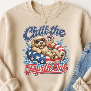 Pode incluir: Uma camisola de gola redonda bege com o gráfico de um cão dourado com óculos de sol vermelhos, a flutuar numa boia com estrelas vermelhas, brancas e azuis enquanto segura uma bebida. O texto diz Chill the Fourth Out em azul e vermelho.