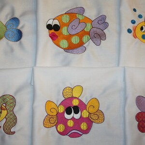 May include: Six embroidered designs of colorful fish on white fabric. The fish have different patterns and colors, including yellow, orange, green, blue, pink, and purple.