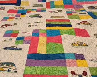 May include: A colorful patchwork quilt featuring embroidered images of vintage campers, cars, bicycles, and camping chairs. The quilt has a white background with floral stitching and blocks of pink, blue, green, and yellow. The words "We Love Camp" are embroidered in the upper left corner.