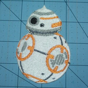 May include: Embroidered patch featuring the Star Wars droid BB-8. The patch is white with orange and gray accents, including circular details and a domed head. The design is detailed with black stitching.