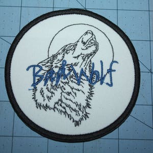 May include: A white embroidered patch with a black and white wolf howling at the moon. The text "Bad Wolf" is embroidered in blue.