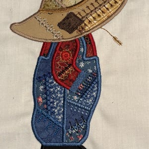 May include: Appliqued fabric art of a figure wearing a tan hat and blue overalls. The hat has a brown patch and decorative stitching. The overalls are blue with floral and geometric patterns, and a red bib. The figure has a black base.