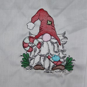 May include: Embroidered design of a gnome holding a candy cane. The gnome is wearing a red hat with a green star and has a white beard. The gnome is surrounded by green leaves.