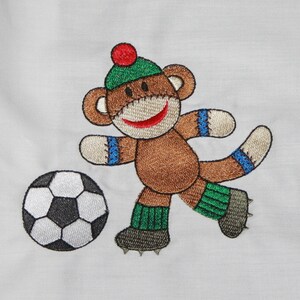May include: Embroidered design of a brown monkey wearing a green hat and blue and green socks, kicking a black and white soccer ball.