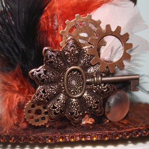 May include: A black, orange, and white top hat with a steampunk design. The hat is decorated with feathers, gears, a key, and a decorative metal piece.