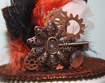May include: A black, orange, and white top hat with a steampunk design. The hat is decorated with feathers, gears, a key, and a decorative metal piece.