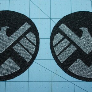 May include: Two black and silver embroidered patches featuring the S.H.I.E.L.D. logo. The logo is a circle with a silver eagle inside.