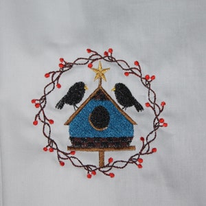 May include: Embroidered design of a blue birdhouse with a gold star on top, surrounded by a wreath of red berries and brown vines. Two black birds are perched on the birdhouse.