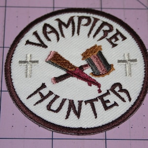 May include: White embroidered patch with a brown border. The patch features a crossed hammer and stake with a red blood drop. The text "Vampire Hunter" is written in a curved arc around the image. Two white crosses are on either side of the image.