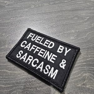 以下が含まれることがあります： 「FUELED BY CAFFEINE & SARCASM」と白い刺繍が施された黒い長方形のパッチ。 パッチには黒い縁取りがあり、衣類やアクセサリーに縫い付けたり、アイロンで接着したりするように設計されています。 ユーモラスなアクセサリー。
