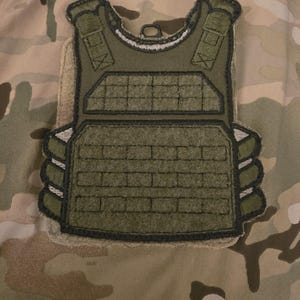May include: A green tactical vest patch with black stitching and a velcro backing. The patch features a stylized depiction of a tactical vest with a front panel and side plates.