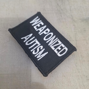 Può includere: Toppa rettangolare nera con testo ricamato bianco che recita "WEAPONIZED AUTISM". La toppa è probabilmente in tessuto e ha un bordo cucito. La toppa è su una superficie grigio chiaro.
