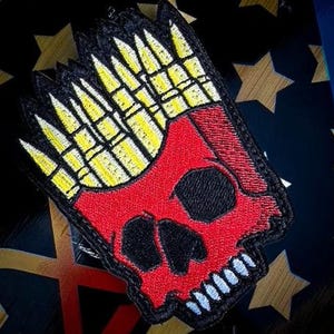 May include: Embroidered patch featuring a red skull with black eye sockets and a white-toothed grin. The skull is topped with yellow and black bullet-shaped designs. The patch has a black border.