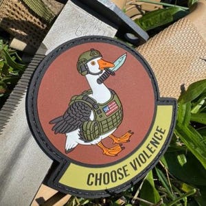 Könnte beinhalten: Runder Aufnäher mit einer Cartoon-Gans, die einen Militärhelm und eine Weste trägt und ein Messer im Schnabel hält. Der Aufnäher hat einen braunen Hintergrund und ein gelbes Banner mit dem Text "CHOOSE VIOLENCE".