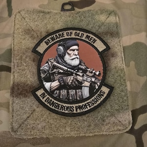 Op de afbeelding: Een beige patch met een zwarte rand en de tekst "Beware of Old Men - A Dangerous Profession" in een cirkelvormig ontwerp. Het midden van de patch toont een cartoonillustratie van een man met een baard en een geweer.
