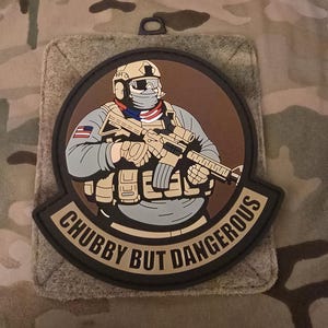 Puede incluir: Un parche redondo negro con fondo marrón. El parche presenta una ilustración de dibujos animados de una persona con casco y sosteniendo un rifle. El texto "Chubby But Dangerous" está escrito en letras doradas alrededor del borde del parche.