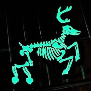 Pode incluir: Um patch preto com um design de esqueleto de veado verde brilhante. O veado é retratado numa pose de corrida, com os seus chifres e estrutura esquelética claramente visíveis. Ideal para roupas ou acessórios.