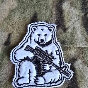 Peut inclure: Patch brodé représentant un ours polaire blanc avec un contour noir tenant un fusil noir.